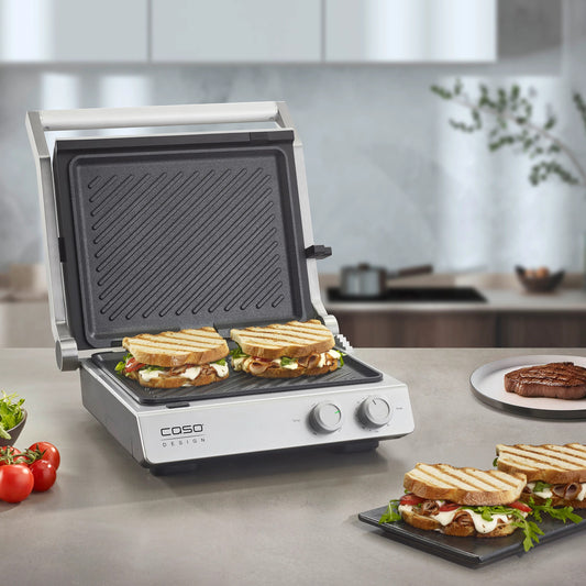 Piastra elettrica SteakChef CASO Design aperta su tavolo da cucina con bistecche in cottura