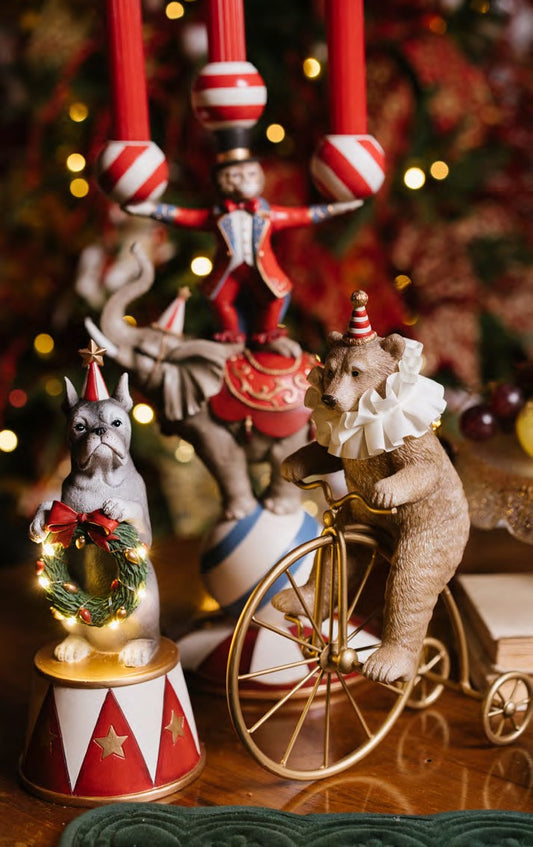 Statuina natalizia decorativa in resina a forma di orsetto marrone chiaro da circo con colletto bianco, mentre pedala su un antico biciclo vintage color oro lucido, altezza 25,5 cm, collezione Velvet Luxury Nuvole di Stoffa, Tutto Chic.