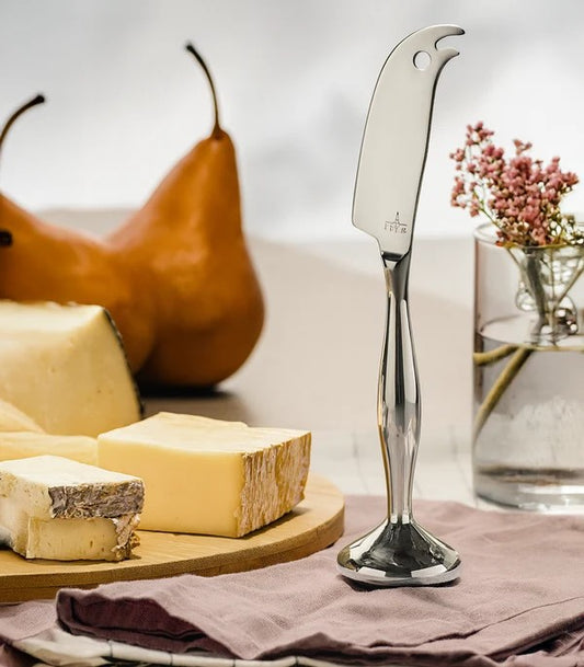 Coltello basculante in acciaio inox I Genietti appoggiato in piedi sulla tovaglia accanto a un tagliere di formaggi e pere