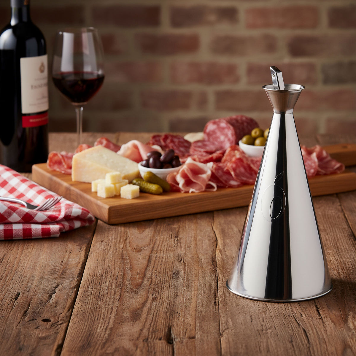 Oliera da tavola Olivella Magnum Olipac in acciaio inox posizionata su tavolo rustico accanto a tagliere di salumi e formaggi